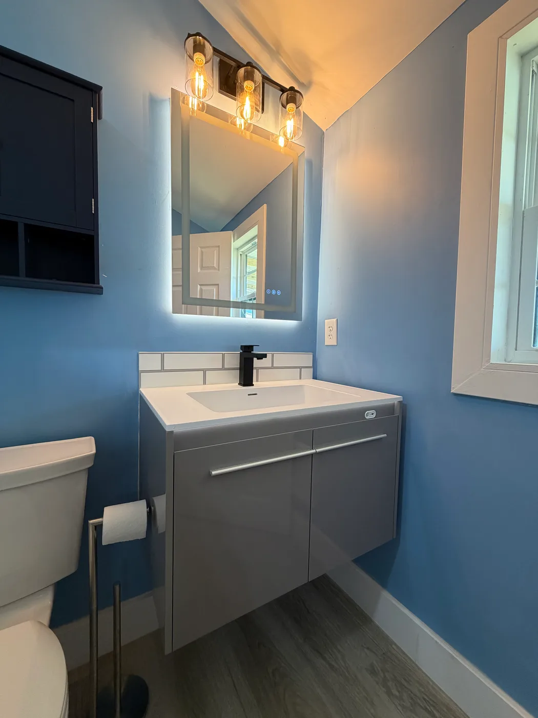 Floating gray vanity with a white vessel sink, black faucet and a backlit LED mirror