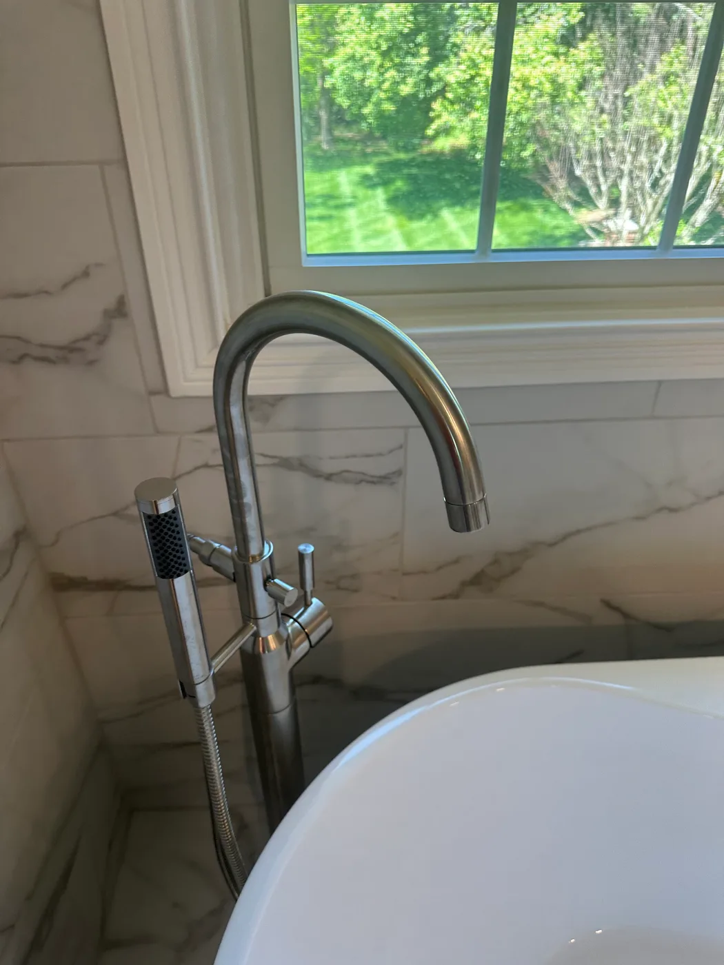 Brushed nickel floor-mount tub filler with a handheld sprayer beside a bright window