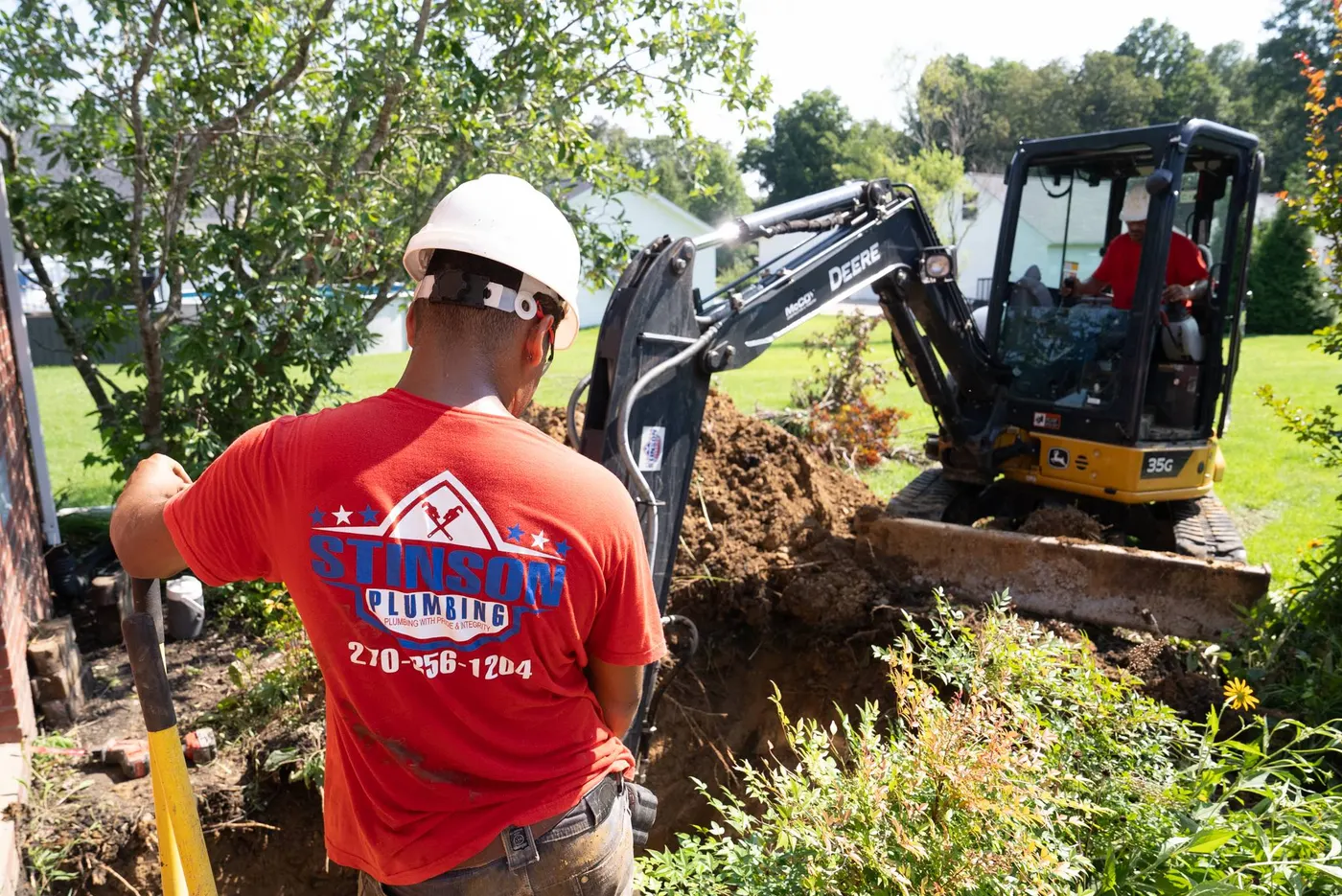 Tech in a red shirt and hard hat with a mini excavator in a residential yard